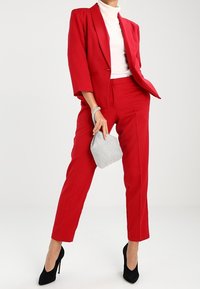Blazer rouge et pantalon assorti en tissu tissé. Le blazer présente un revers cranté, et le modèle tient une pochette à carreaux et porte des talons noirs.