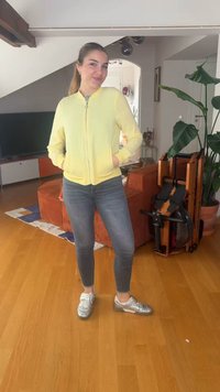 Νεαρή γυναίκα με κίτρινο μπουφάν και γκρι τζιν στέκεται χαμογελώντας σε ένα σαλόνι με ξύλινο πάτωμα, πορτοκαλί καναπέ και φυτά εσωτερικού χώρου.