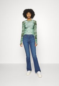 Top de manga larga con un patrón de espiral verde y blanco, con mangas transparentes, combinado con vaqueros acampanados azules y zapatillas blancas. Diseño sencillo.