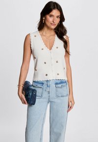 Top senza maniche bianco in maglia con scollatura a V, rifiniture in pizzo e decorazioni floreali, abbinato a jeans larghi azzurri e una borsa blu.