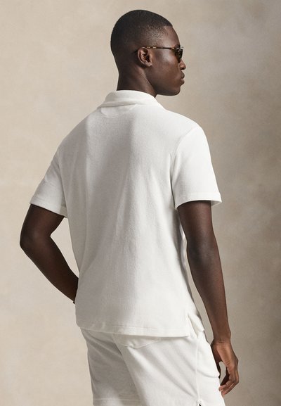 Λευκό πόλο πουκάμισο με υφή, κοντά μανίκια και επίπεδη γιακά, με πλευρικές χαραμάδες για ευκολία στην κίνηση, φορεμένο με ταιριαστά σορτς.