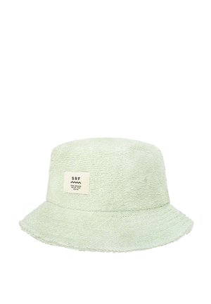 Kobieta poprawia beżową kapelusz wiadro, nosi biały top bikini, pasujące szorty i gumkę do włosów, stoi obok drzewa z zielonymi liśćmi i białymi kwiatami.