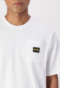 White cotton t-shirt featuring a round neckline and a chest pocket with a black label displaying "StunRay" in golden text.