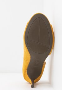 Żółty zamszowy but na wysokim obcasie z wąskim czubkiem i smukłym obcasem typu stiletto. Podeszwa jest ciemnobrązowa z teksturowanym wzorem antypoślizgowym.