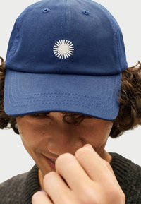 Blauwe pet met een gebogen rand, voorzien van een witte zonnestraal borduurwerk in het midden. Gemaakt van textuurstof met gestikte accenten.