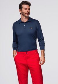 Polo a maniche lunghe in blu navy con tre bottoni e un logo, abbinato a pantaloni rossi brillanti. Il tessuto sembra liscio con un design aderente.