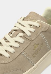 Beige Wildledersneaker mit weißen Schnürsenkeln, goldenem Logo und dekorativen Nähten entlang der Sohle. Verfügt über eine glatte Textur und abgerundete Form.