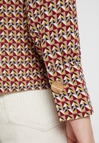 Gros plan sur le poignet d'une chemise à manches longues à motif avec un bouton métallique rond porté par-dessus un pantalon blanc texturé.