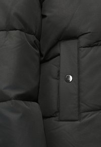 Tuvumā redzama melna quiltota jaka kabata ar vertikālu spiedpogu un klātām auduma plāksnēm.