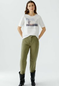 Weißes Baumwoll-T-Shirt mit einem Grafikdesign eines Wüsten-Sonnenuntergangs, kombiniert mit olivgrünen, taillierten Hosen und schwarzen knielangen Stiefeln.