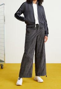 Tmavě šedá zipová bunda s bílými detaily, v kombinaci s širokými texturovanými kalhotami a bílými teniskami. Modelka stojí na žluté podlaze.