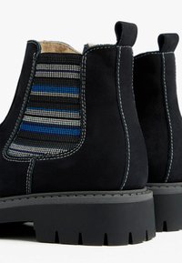 Bottines noires en daim avec un panneau élastique rayé texturé en bleu, gris et blanc, dotées d'une semelle en caoutchouc noire robuste.