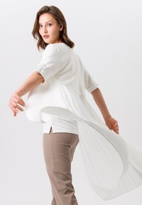 Weiße Bluse mit schichtweisem Design und gefaltetem Rückendetail, kombiniert mit beigen Hosen. Die Bluse hat kurze Ärmel und einen hohen, asymmetrischen Saum.