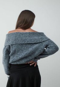 Grijze off-the-shoulder trui met geribbelde halslijn en textuur breiwerk. Beschikt over lange mouwen en een relaxte pasvorm, gecombineerd met een zwarte rok.