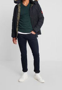 Man in een donkere pufferjack met een bontkap, groene V-hals trui, donkere spijkerbroek, witte sneakers, staande met één hand in zijn zak op een effen achtergrond.