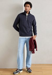 Felpa blu navy con colletto alto, apertura a mezza zip; abbinata a jeans azzurri e sneakers multicolori, tiene una felpa bordeaux.