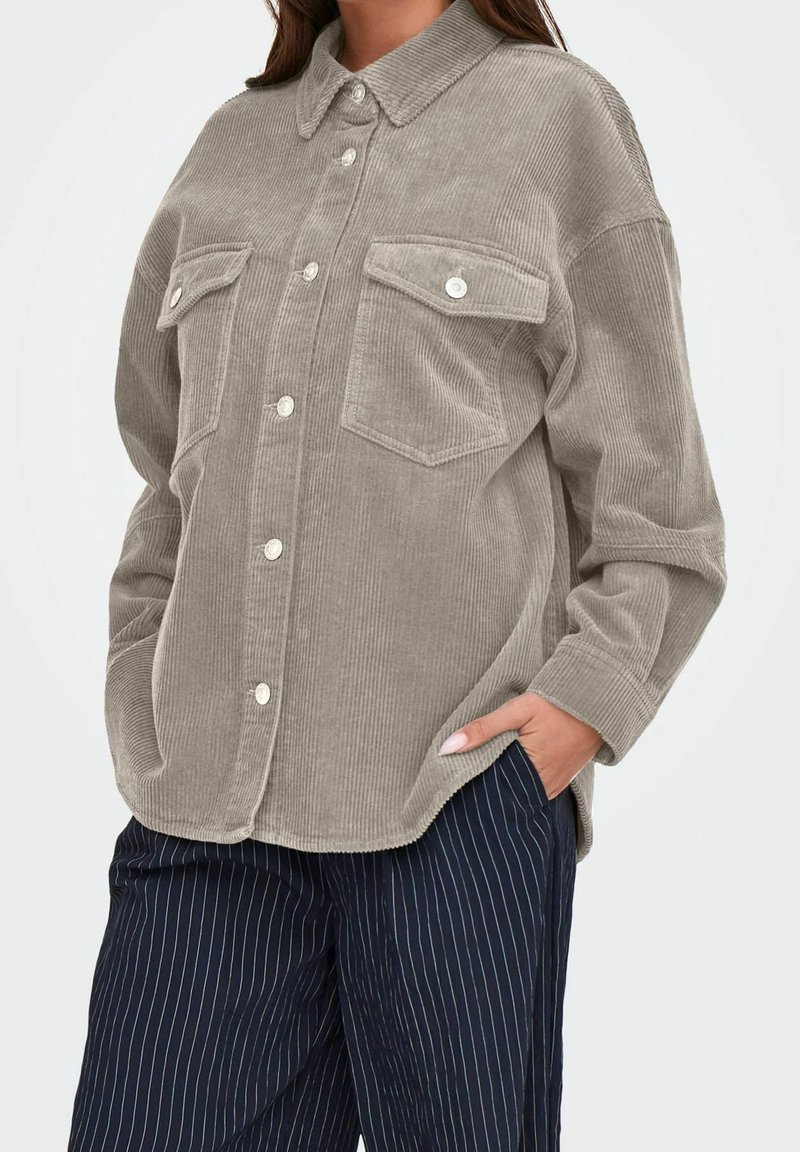 Femme portant une chemise en velours côtelé grise boutonnée avec deux poches poitrine et un pantalon bleu marine à fines rayures, les mains partiellement dans les poches.