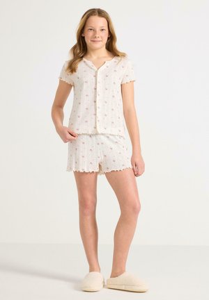 Meisje met lichtbruin haar, gekleed in een crèmekleurig shirt met korte mouwen en bloemmotief, bijpassende korte broek en crèmekleurige slippers, staand tegen een witte achtergrond.