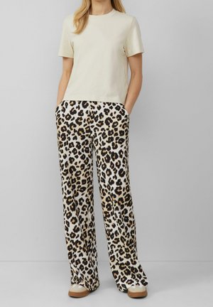 Vrouw die een crème-kleurige korte mouw top en brede broek met luipaardprint draagt, met handen in de zakken, staand tegen een effen grijze achtergrond.