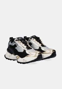 Par de zapatillas gruesas en negro, blanco y dorado con cordones en la parte frontal, suelas texturizadas y detalles metálicos sobre un fondo blanco.