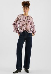 Przewiewna, półprzezroczysta różowa bluzka z wzorem tie-dye i falbanianymi rękawami, w połączeniu z ciemnoniebieskimi szerokimi spodniami dżinsowymi oraz czarnymi botkami za kostkę.