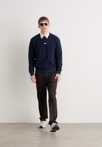 Marine genser med ribbestrikkede mansjetter og kant, hvit skjorte under, brune bukser og grå joggesko. Modell som har på seg solbriller.