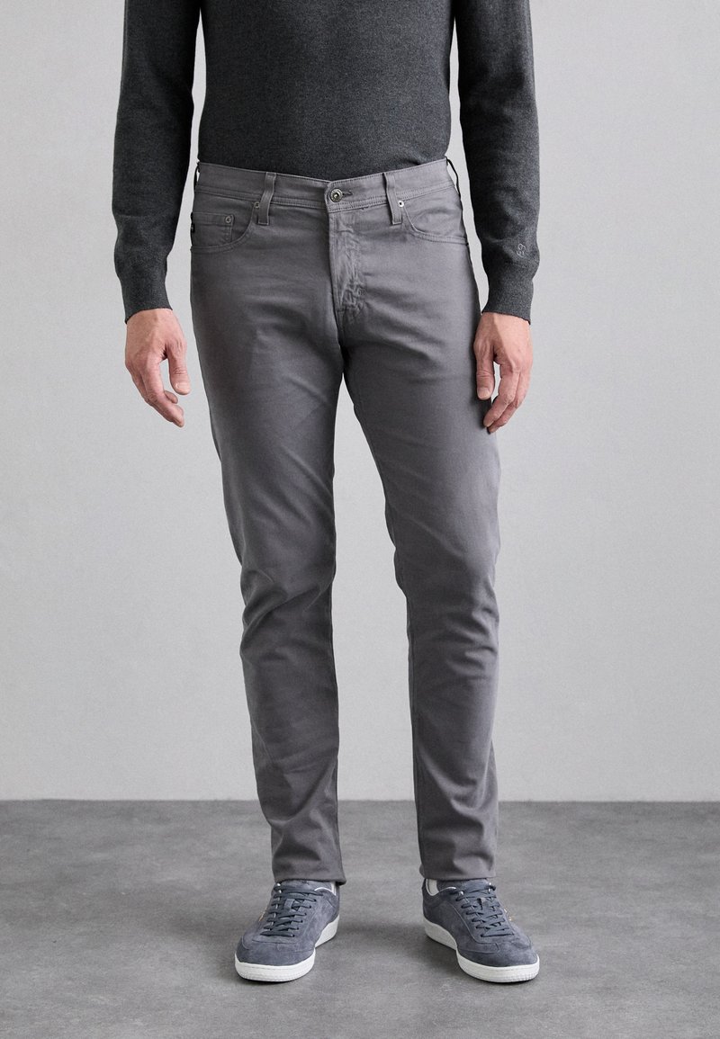 Pantalones ajustados gris con cinco bolsillos, cierre frontal con botón y bucles para cinturón. Combinados con zapatillas gris y un suéter oscuro.
