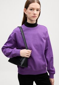 Felpa viola con un sottogiacca nero a coste sotto. Il modello tiene in mano una piccola borsa nera. Design semplice, texture morbida.