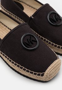 Espadrilles en tissu noir avec des semelles tissées beige et un logo circulaire noir avec les lettres MK sur le dessus.