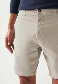 Bermudas beige de tela ligera con un frente plano. Presenta un cierre de botón y bolsillos laterales. Textura suave con costuras sutiles.