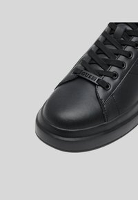 Svarte skinnsneakers med rund tå, strukturert overflate, avrundet såle og fremtredende tungelapp med "GUESS"-logo i svart.