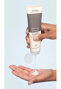 Aveda Damage Remedy dagelijkse haarherstel tube, grijs en crème, die een witte crème in een hand dispent. De tube heeft een flip-top dop.