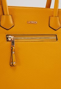 Sac à main en cuir texturé jaune avec poche zippée dorée et plaque de marque assortie dorée à l'avant.