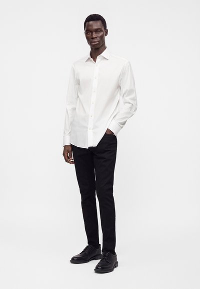 Uomo in piedi con una mano in tasca, indossa una camicia bianca con bottoni, pantaloni neri e scarpe eleganti nere su uno sfondo semplice.