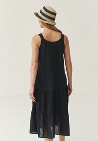 Black tiered cotton dress with spaghetti straps and a flowing silhouette. Paired with a wide-brimmed striped straw hat.