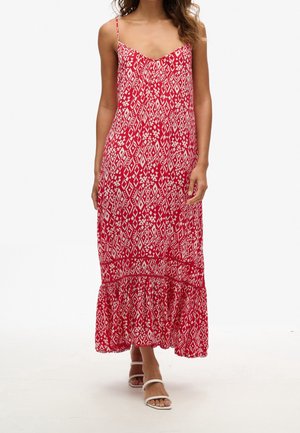 Kvinde iført en rød- og hvidmønstret ærmeløs maxikjole med tynde stropper og hvide åbne sandaler, stående på en hvid baggrund.