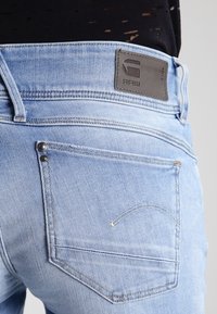 Svetle modré džíny z denimu s vybledlým omakem, které mají na pásku černou koženou nášivku s nápisem "RAW" a standardní zadní kapsy.