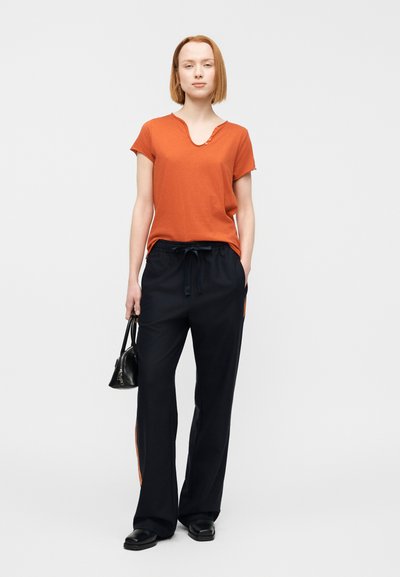 Πορτοκαλί κοντομάνικο μπλουζάκι με V-λαιμόκοψη και λεπτομέρεια κουμπιών, σε συνδυασμό με φαρδιά μαύρη φόρμα με πορτοκαλί ρίγες στο πλάι. Μαύρη τσάντα χειρός.