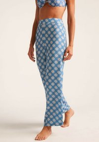 Mujer con pantalones de tiro alto azules y un top de bikini a juego con un patrón floral blanco, de pie descalza sobre un fondo claro.