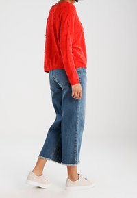 Roter strukturierter Pullover mit langen Ärmeln, kombiniert mit blauen, cropped Jeans mit fransigen Säumen und beigen Sneakers. Lässiges Outfit.