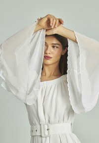 Weißes Kleid mit fließenden, gewellten Glockenärmeln, gerafftem Taillenbereich und einem breiten Gürtel. Leichte Stoffe, glatte Textur, minimalistisches Design.