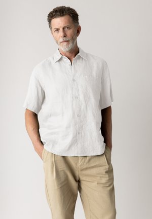 Reifer Mann mit grauem Bart, der ein weißes kurzes Hemd mit Knöpfen und beigen Hosen trägt, steht mit den Händen in den Taschen vor einem schlichten Hintergrund.