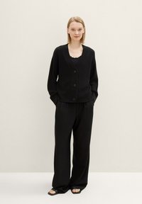 Svart ribbestrikket cardigan med V-hals, frontknapper og lange ermer, parret med svarte vide bukser og flate sandaler.