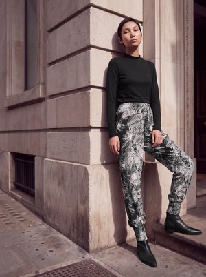 Une femme porte un haut noir, un pantalon à motifs et des bottines. Appuyée contre un bâtiment en pierre, elle pose un pied sur une marche.