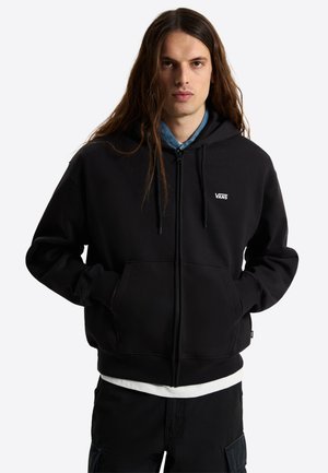 Giovane uomo con capelli lunghi indossa una felpa nera Vans con zip sopra una camicia blu e una maglietta bianca, mani nelle tasche davanti, su uno sfondo semplice.