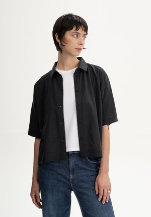 Jonge vrouw met kort donker haar, gekleed in een zwart overhemd met knopen over een wit shirt en blauwe jeans, kijkt opzij tegen een effen achtergrond.