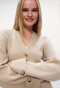 Beige cardigan med V-hals, med store, runde knapper og brede ermer. Myk tekstur og minimalistisk design.