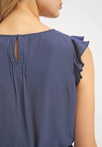 Top azul marino con mangas cortas con volantes, abertura en la parte trasera en forma de ojo de cerradura y una textura de tela suave y lisa. Diseño casual con un corte holgado.