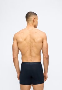 Μυώδης άνδρας που στέκεται με την πλάτη, φορώντας σκουρόχρωμα στενά ανδρικά εσώρουχα, μπροστά σε απλό λευκό φόντο.