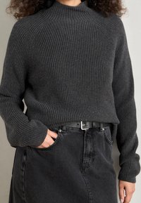 Personne portant un pull en maille côtelée gris foncé rentré dans une jupe en denim noire avec une ceinture noire, main dans la poche de la jupe, sur un fond neutre.